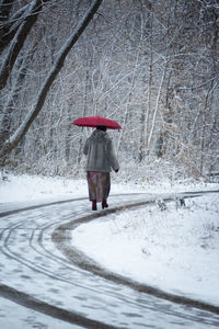 Woman in a gray fur coat with a red umbrella walks through a snow-covered park.