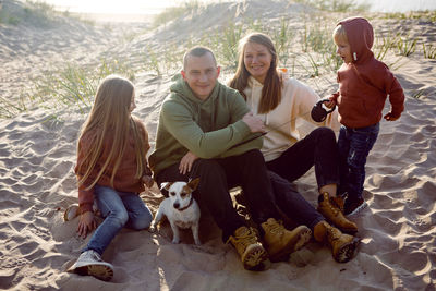 Family with a son and daughter and a dog sit on the sand in autumn