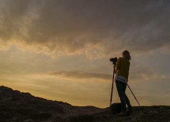 Low angle view of female photographer standing by tripod against cloudy sky during sunset