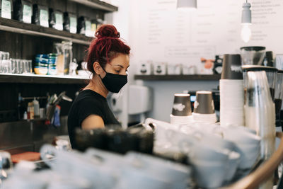 Side view of female bartender in face mask standing at counter in coffee shop and making delicious hot beverage in coffee machine during coronavirus epidemic