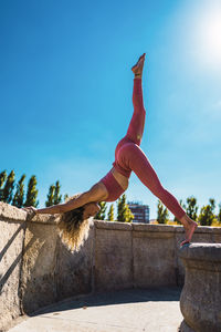 Full body side view of slim female in sportswear doing three legged downward facing dog asana with hands and leg on stone borders while practicing yoga on terrace in sunny day