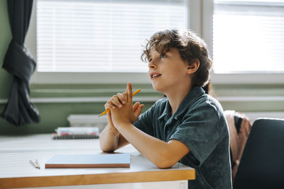Elementary schoolboy with hands clasped sitting near desk in classroom at school