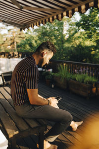Full length of man using smart phone while sitting on bench at back yard