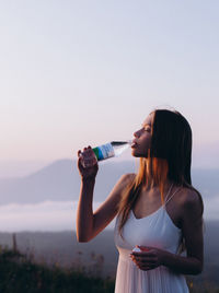 Young woman drinking water from bottle while standing on land against sky