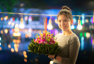Portrait of smiling woman holding flowers while standing against illuminated lights