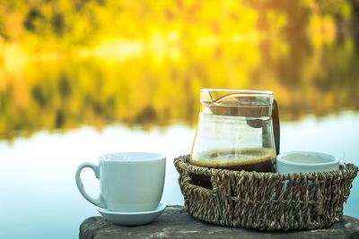 Close-up of coffee in jug by cups on rock against lake