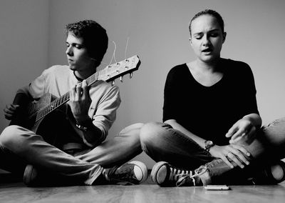 Young man with guitar by friend sitting on hardwood floor at home