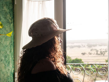 Woman wearing hat while looking through window at home