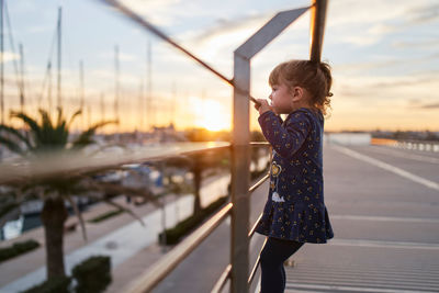 Side view of curious little girl in casual wear standing near railing on urban bridge and looking away in summer evening