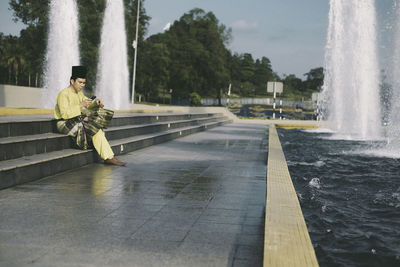 Full length of man in traditional clothing using phone on steps by fountain