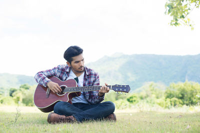 Man playing guitar while sitting on field against sky