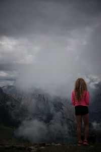 Rear view of woman standing at peak while looking at mountains in foggy weather