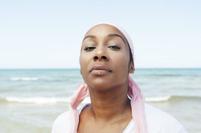 Determined ethnic female with headdress and piercing standing on beach on background of sea and looking at camera