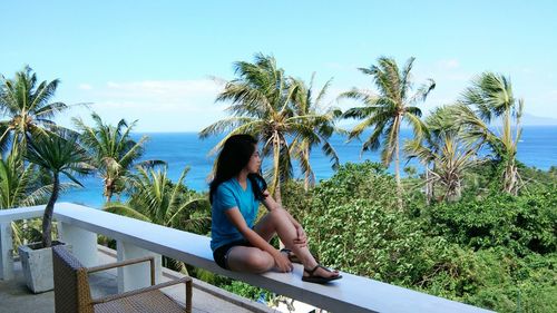 Young woman sitting on balcony by trees and sea against sky