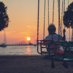 Rear view of boy sitting on swing at beach against sky during sunset