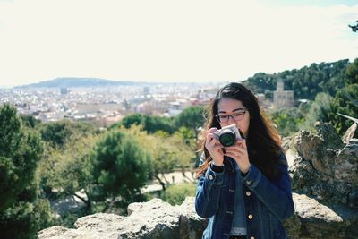 Portrait of young woman photographing against sky