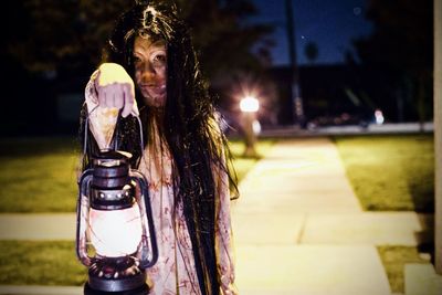 Portrait of girl holding lantern standing on footpath at night