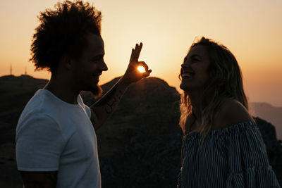 Side view of smiling hipster showing ok against sunbeams and looking at trendy woman laughing while standing against mountain ridge under colorful sky during sunset