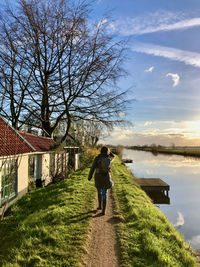 Rear view of a woman walking on a small dike along a canal, a farmhouse and bare trees