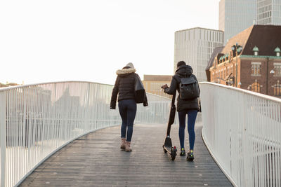 Rear view of female friends walking with e-scooter on bridge in city