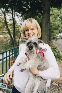 Portrait of smiling blond woman holding cute dog in public park