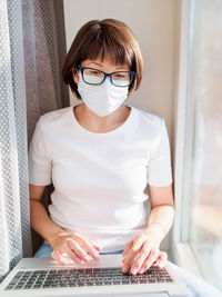 Woman in medical mask remote works from home. she sits on window sill with laptop on knees.