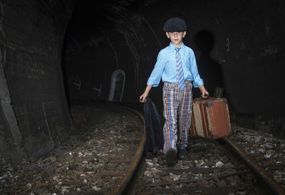 Portrait of boy holding suitcase and walking on railroad track in tunnel 