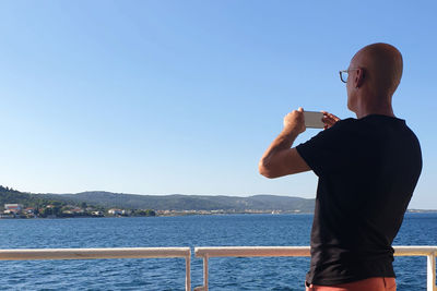 Man standing on ferry boat holding smartphone taking picture of island