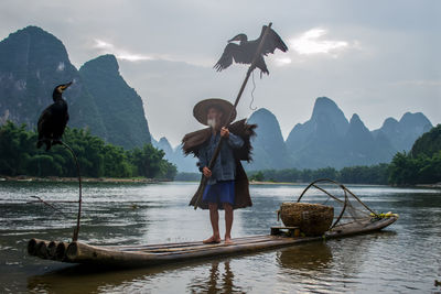 Cormorants flying over man standing in boat on river against mountains