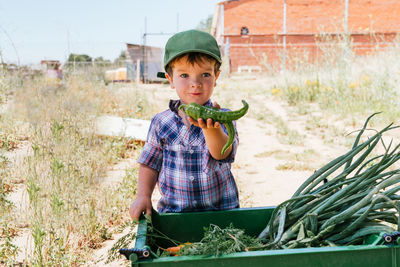 Close-up of a little blond boy in a cap and plaid shirt showing a pepper he has picked in his garden