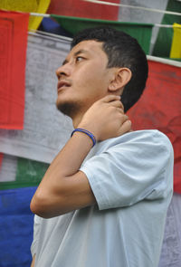 A asian guy posing with hand behind neck looking sideways, standing against buddhist prayer flags  