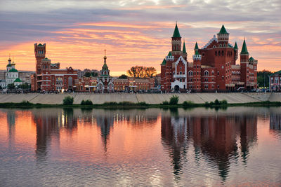 Buildings by river against sky during sunset