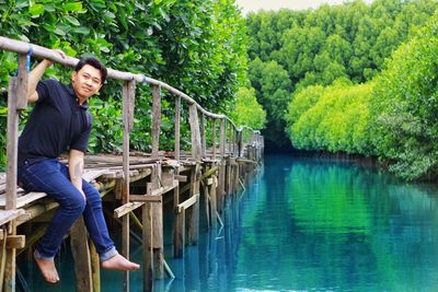 Portrait of smiling young man sitting on pier over lake in forest