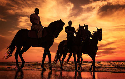 Silhouette people riding horse on shore against sunset sky