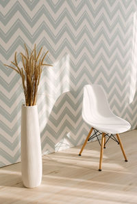 A white vase with decorative dried plants next to a white chair in the stylish interior of the room