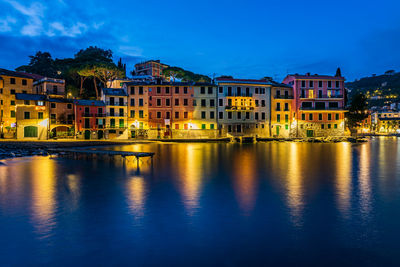 Illuminated buildings by river against sky at dusk