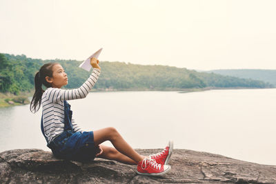 Side view of girl playing with paper airplane while sitting on rock against lake
