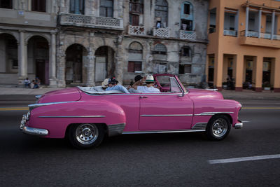 People traveling in pink vintage car on city street against buildings