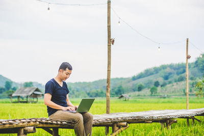 Man using laptop while sitting on boardwalk at rice paddy