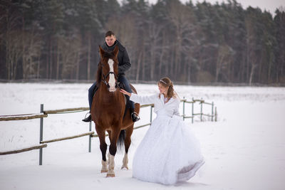 Groom sitting on horse by bride on snow covered field