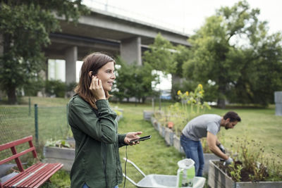 Mid adult woman talking on mobile phone with man planting in background at urban garden