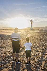 Rear view of man with son standing on beach against lighthouse