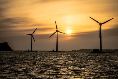 Silhouette windmills by sea against sky during sunset