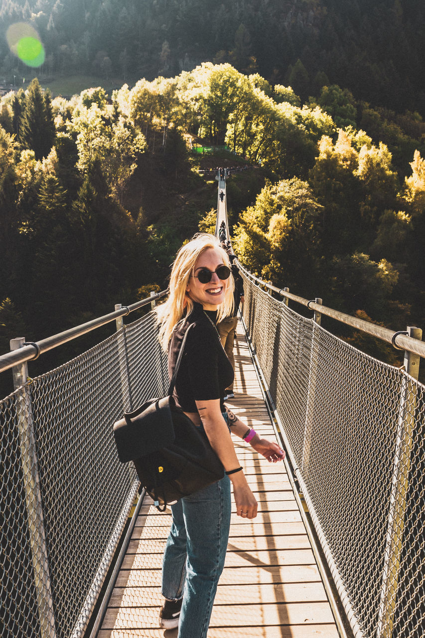 one person, real people, young adult, young women, railing, portrait, standing, leisure activity, casual clothing, lifestyles, connection, tree, front view, glasses, nature, plant, looking at camera, architecture, hair, fashion, hairstyle, outdoors, footbridge