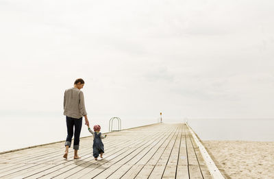 Rear view of mother and daughter walking on pier at beach against sky