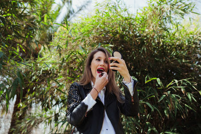 Mid adult woman applying lipstick while standing against plants in park