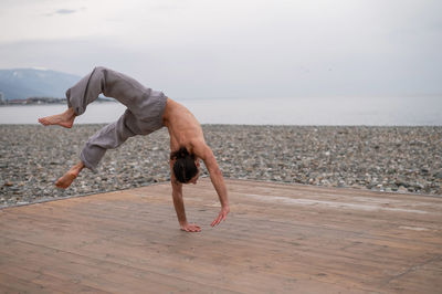 Shirtless caucasian man doing backflip on pebble beach