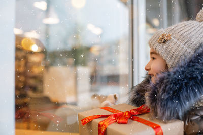 Cute smiling girl with christmas gift looking through window