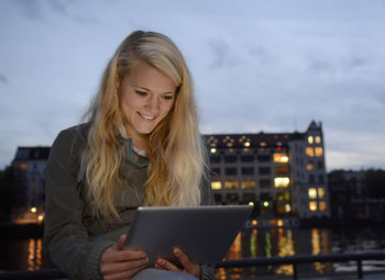 Young woman using digital tablet while sitting in city against sky