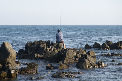 Rear view of man fishing while sitting on rock by sea against clear sky